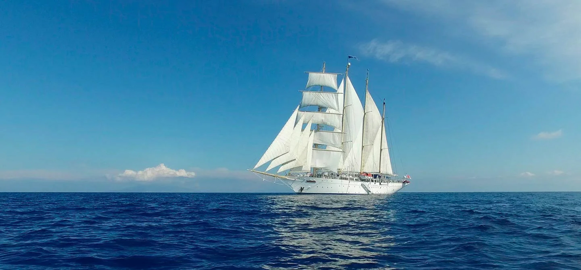 White Sailing Ship on Blue Ocean A classic three-masted tall ship with fully unfurled white sails cruising on a pristine blue ocean under a clear sky. The elegant vessel appears to be a modern sailing yacht or training ship.