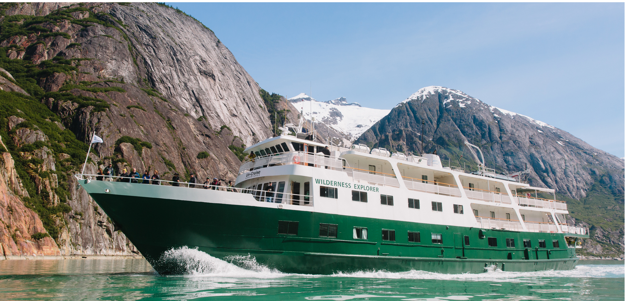 Wilderness Explorer Cruise in Alaska A green and white expedition cruise ship navigating through a scenic Alaskan fjord with dramatic rocky mountains and snow-capped peaks in the background.