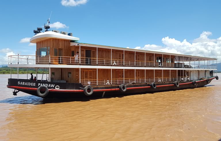 Wooden River Cruise Boat on Muddy Water A two-deck wooden river cruise boat named 'Sabaidee Pandaw' sailing on a muddy brown river with a clear blue sky and white clouds in the background.