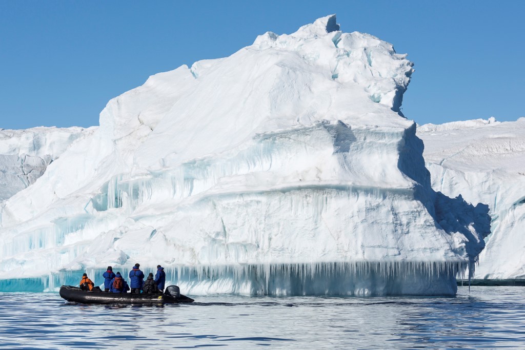 Zodiac Expedition Near Massive Iceberg A small zodiac boat carrying adventurers approaches a towering, pristine white iceberg with distinctive blue ice formations and icicle curtains hanging from its base in Arctic waters under clear blue skies.