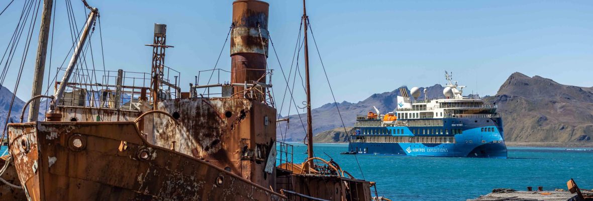 Abandoned Ship and Cruise Vessel in Grytviken A rusted, abandoned whaling ship sits in the foreground of Grytviken harbor in South Georgia, while a modern expedition cruise ship anchors in the turquoise waters behind it, with snow-capped mountains visible across the bay. The stark contrast highlights the region's maritime history and contemporary polar exploration tourism.
