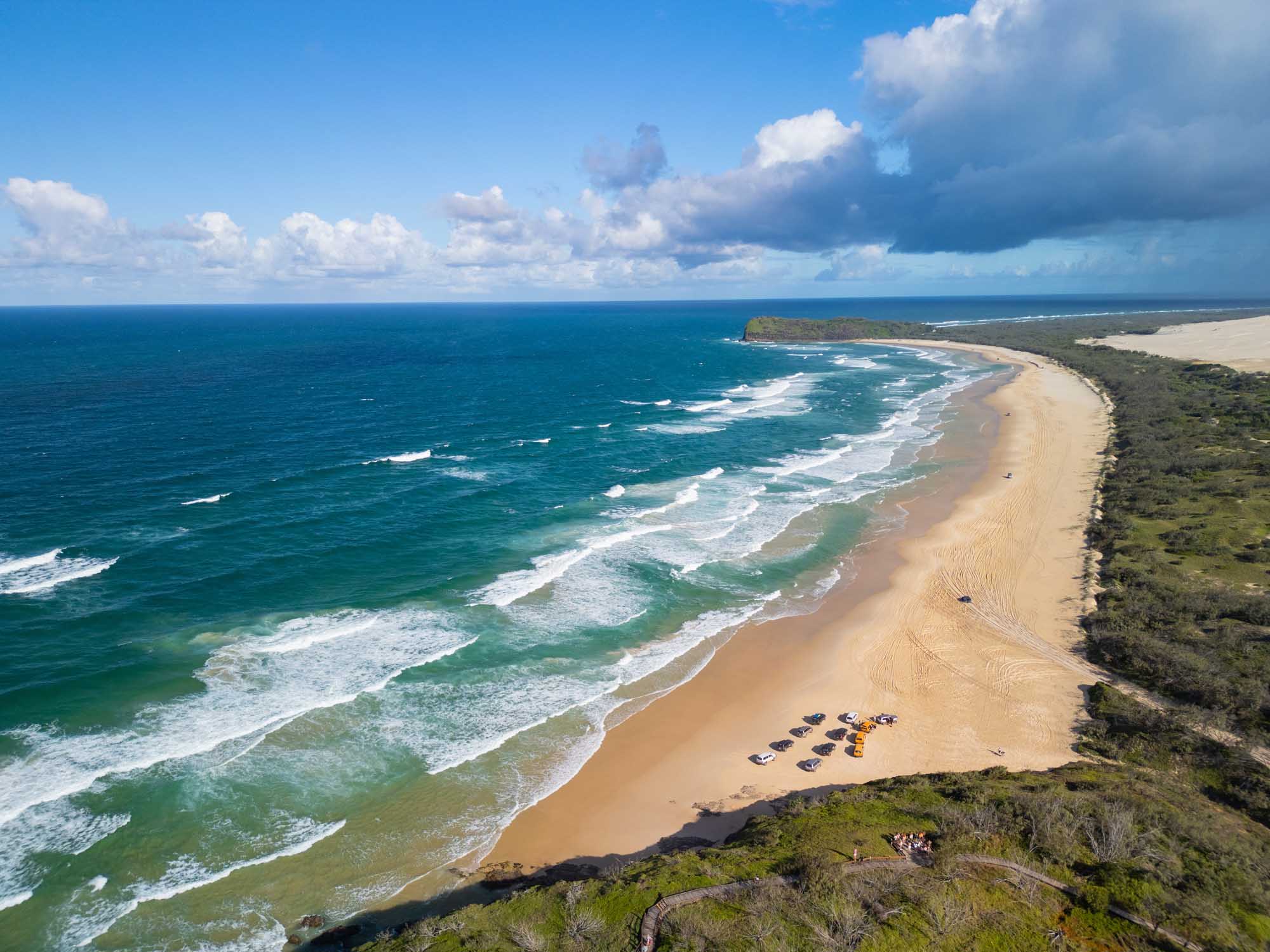 Aerial View of 75 Mile Beach, K’gari Island A stunning aerial photograph of 75 Mile Beach on K'gari Island (Fraser Island) in Queensland, Australia, showing a pristine golden sand beach stretching along turquoise ocean waters with white-capped waves, parked vehicles on the beach, lush green vegetation inland, and a dramatic sky with cumulus clouds.