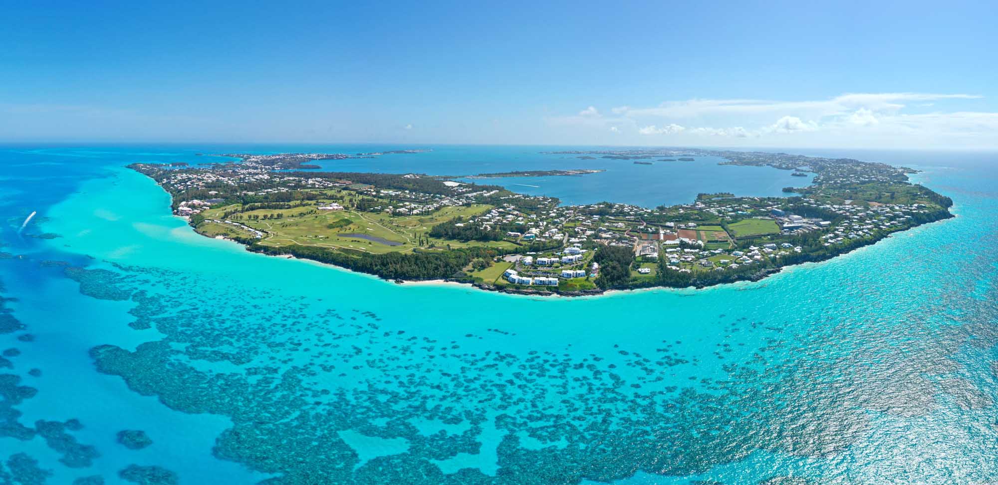 An aerial photograph of Bermuda island surrounded by stunning turquoise Atlantic Ocean waters, showcasing the island's lush green vegetation, residential areas, and pristine beaches. The image captures the island's distinctive crescent shape with clear views of the shallow turquoise waters and darker blue deeper ocean.