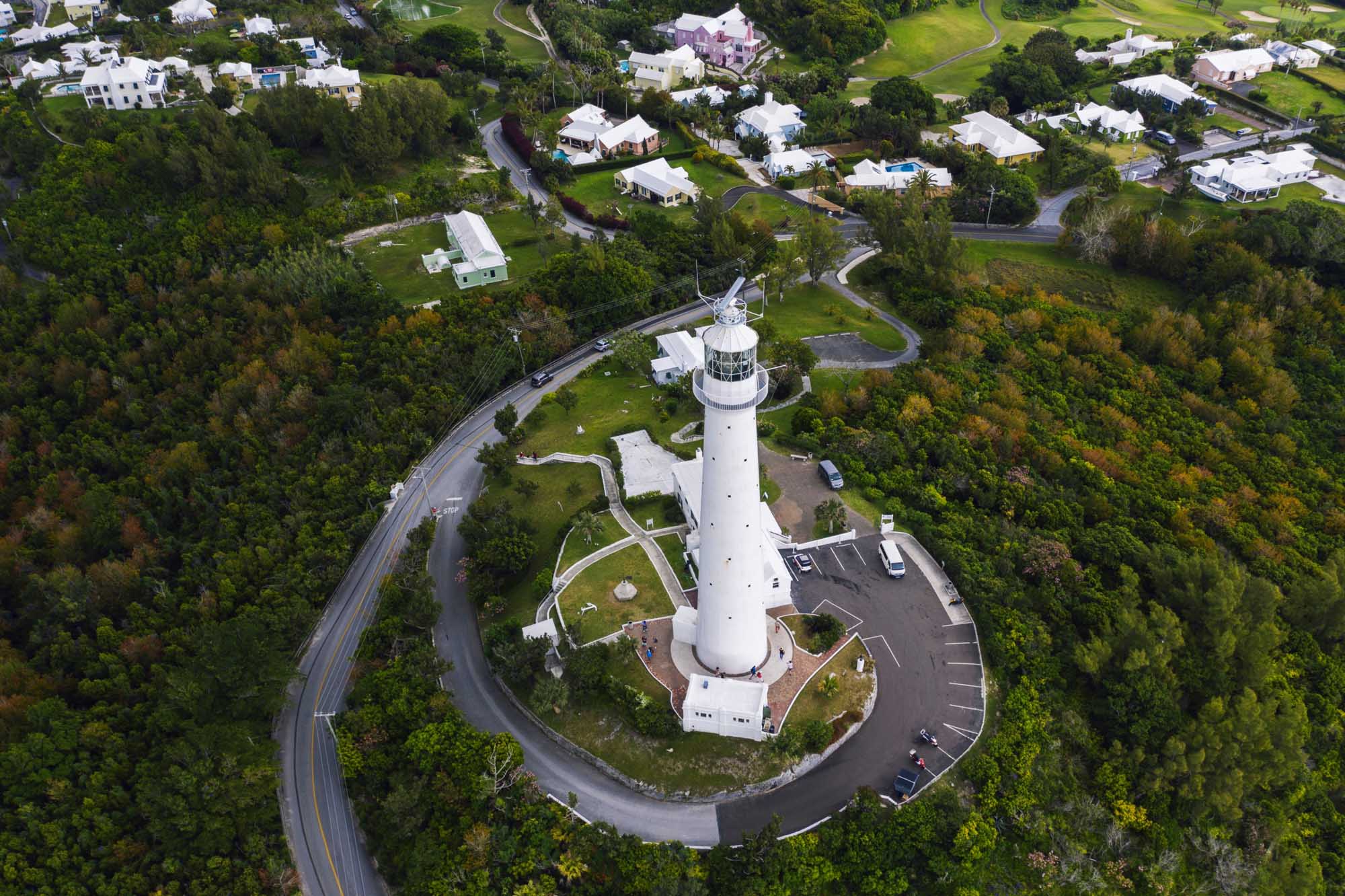 An aerial photograph of a distinctive white lighthouse with its accompanying keeper's residence situated on a promontory in Bermuda, surrounded by lush green vegetation, winding coastal roads, and residential properties. The image showcases the island's verdant landscape, curving waterways, and well-maintained suburban homes characteristic of Bermuda's affluent communities.