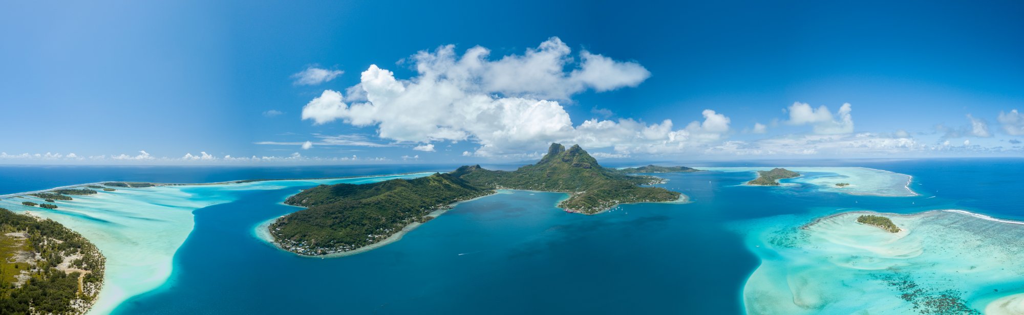 Aerial View of Bora Bora Lagoon and Islands A stunning aerial panorama of Bora Bora, French Polynesia, showcasing the dramatic twin volcanic peaks rising from lush green vegetation, surrounded by crystal-clear turquoise lagoon waters with vibrant coral reef patterns visible in the shallow areas, dotted with small islands and settlements.