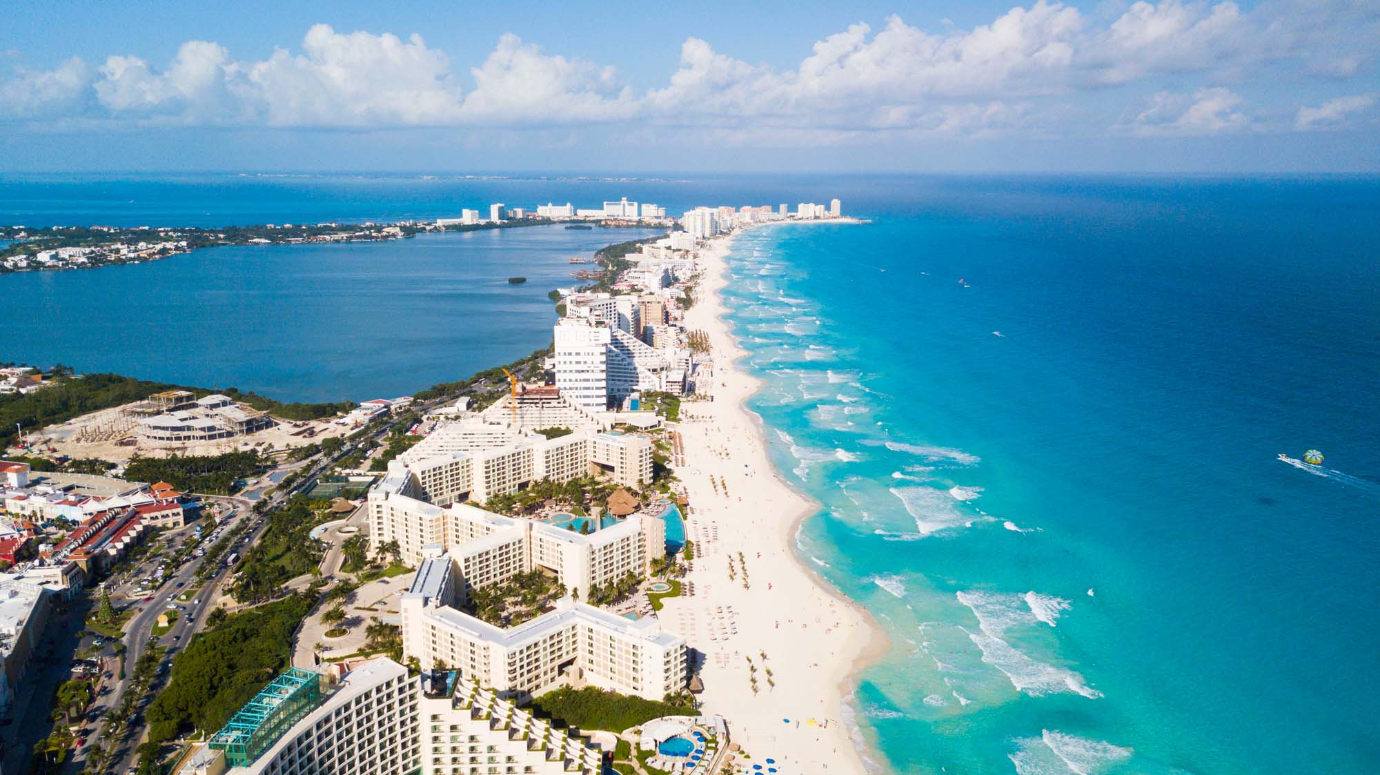 An aerial photograph of Cancun's famous hotel zone showing a narrow strip of white sand beach lined with high-rise resort hotels, turquoise Caribbean waters, and the lagoon on the opposite side. The image captures the distinctive crescent shape of the developed tourist area with clear blue skies and calm waters.