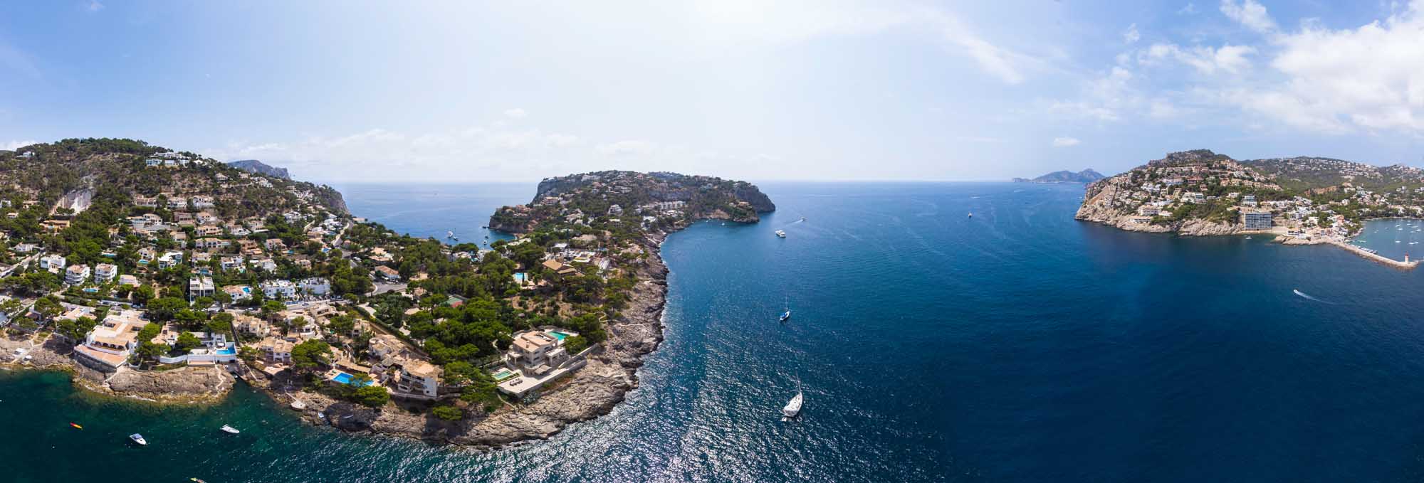 A stunning aerial photograph of Port d'Andratx in Mallorca, Balearic Islands, showcasing dramatic rocky coves, crystal-clear turquoise waters, luxury Mediterranean villas nestled on hillsides, and numerous moored yachts. The natural harbor is surrounded by lush vegetation and rocky peninsulas, with smaller neighboring islands visible in the distance.