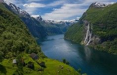 Alpine Fjord with Waterfall and Mountain Peak A stunning alpine fjord landscape featuring a pristine turquoise-blue lake surrounded by steep green mountains, with a dramatic waterfall cascading down the cliff face on the right side and snow-capped peaks visible in the distance.