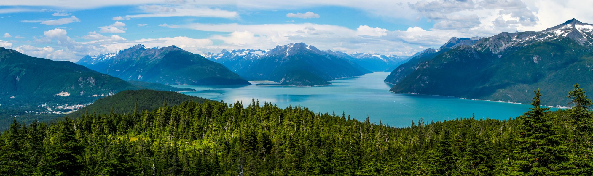 A breathtaking panoramic view of a pristine turquoise alpine lake surrounded by dense coniferous forests and dramatic snow-capped mountain peaks. The scene captures a serene landscape with a small village nestled in the valley below, showcasing the natural beauty of a mountainous lake region in Central Europe.