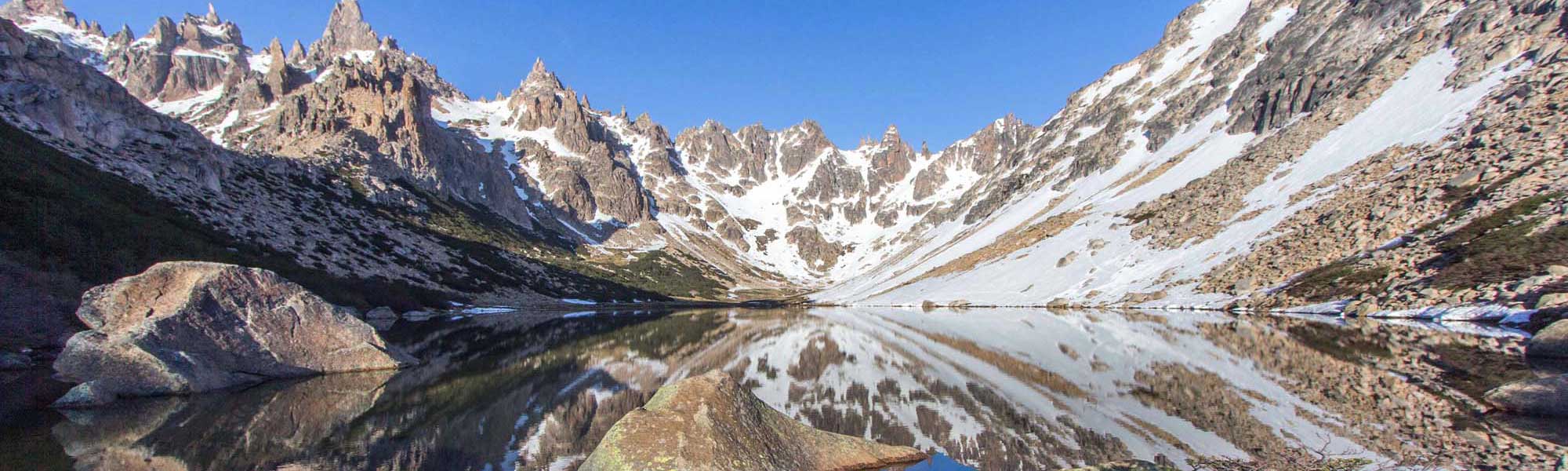 Alpine Lake with Mountain Reflections A pristine high-altitude alpine lake mirrors the dramatic jagged peaks and snow-covered slopes of a mountain range, with clear blue sky and scattered patches of snow visible along the rocky terrain.