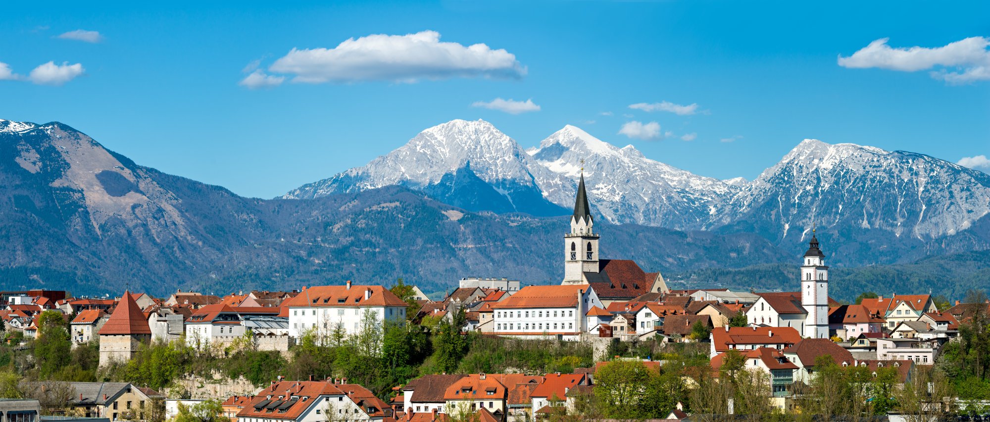 Alpine Village with Snow-Capped Mountain Peaks A picturesque Alpine village nestled in a valley with distinctive orange-tiled roofs and white church spires, framed by dramatic snow-covered mountain peaks in the background under a clear blue sky. The landscape exemplifies the natural beauty and architectural charm of the Bavarian Alps region.