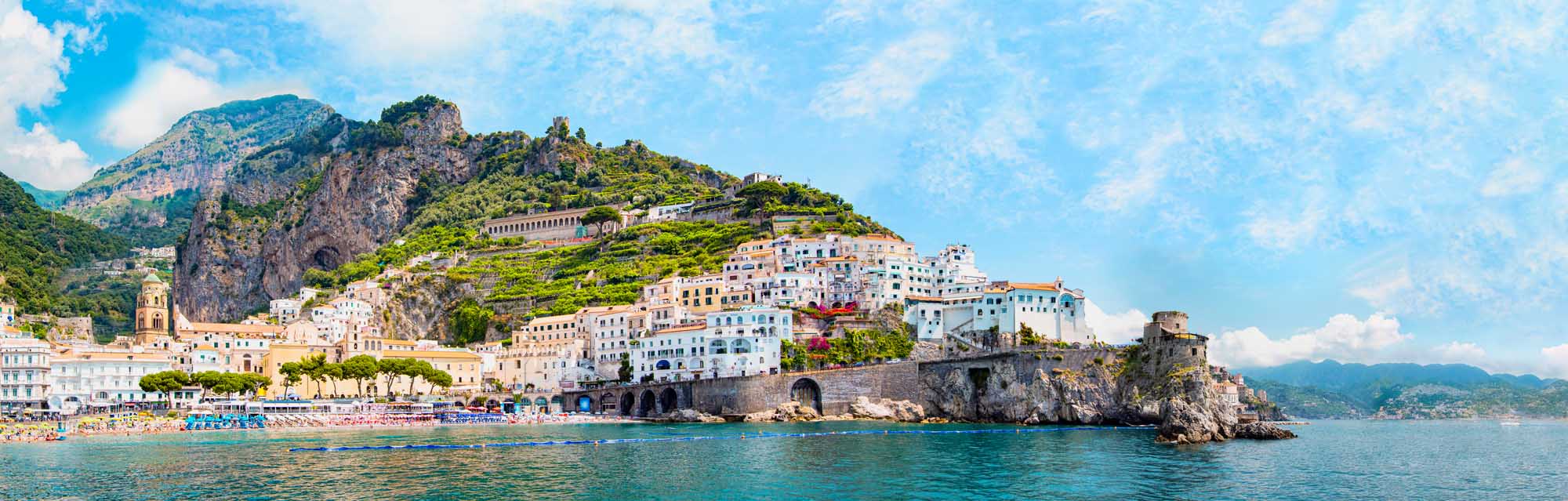 A stunning aerial panoramic view of the picturesque Amalfi village perched on dramatic rocky cliffs along the Amalfi Coast in Italy. The scene showcases colorful pastel-hued buildings cascading down the mountainside, a small sandy beach below, turquoise Mediterranean waters, and lush green vegetation covering the surrounding peaks under a bright blue sky.
