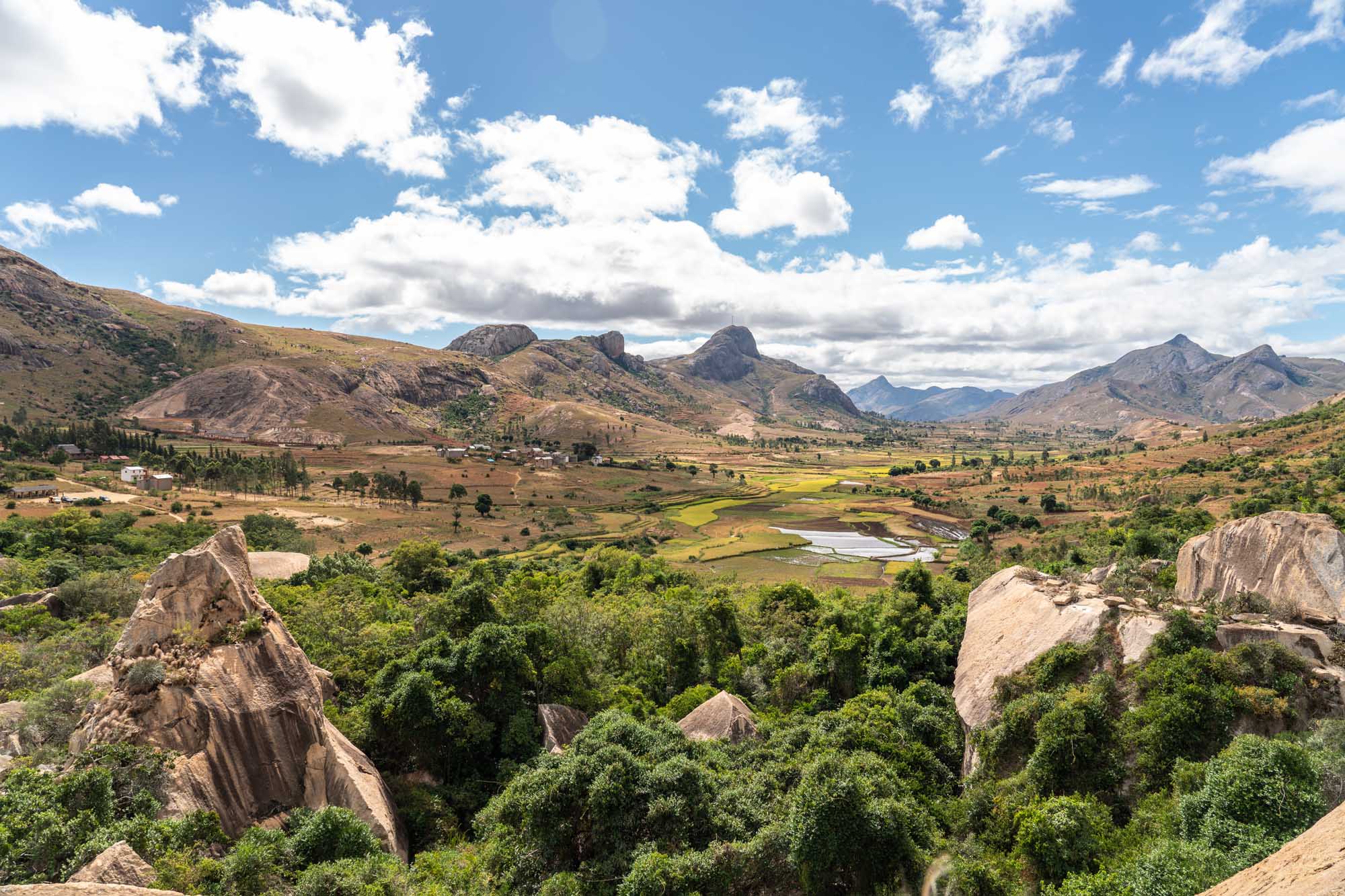 Anja Community Reserve Landscape, Madagascar A panoramic view of the Anja Community Reserve in Madagascar, featuring distinctive large granite boulders scattered across a lush green landscape with rice paddies, distant mountains, and a winding river. The scene combines dramatic rocky outcrops with cultivated agricultural fields and natural vegetation under a partly cloudy sky.