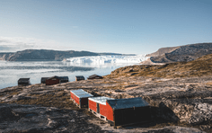 A rustic orange-red cabin sits on a rocky Arctic shore overlooking a pristine fjord filled with icebergs and glaciers in the distance. The stark, barren landscape and massive ice formations exemplify the raw beauty of Arctic exploration and adventure travel.