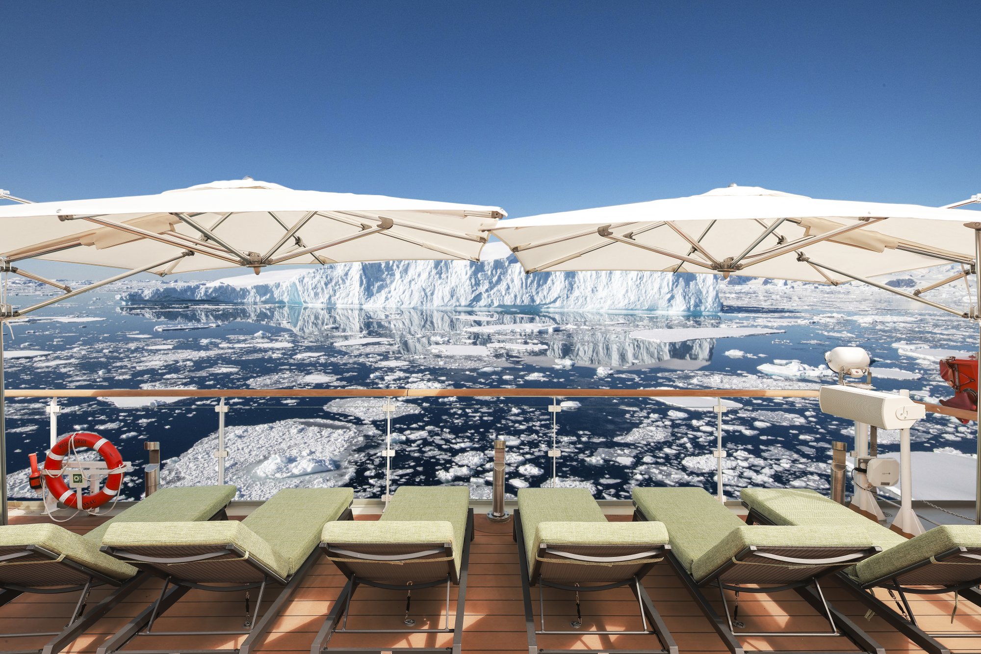 A luxurious sun deck aboard an expedition cruise ship featuring comfortable loungers with green cushions under cream umbrellas, overlooking a pristine Arctic seascape dotted with icebergs and a massive glacier calving wall in the distance under clear blue skies.