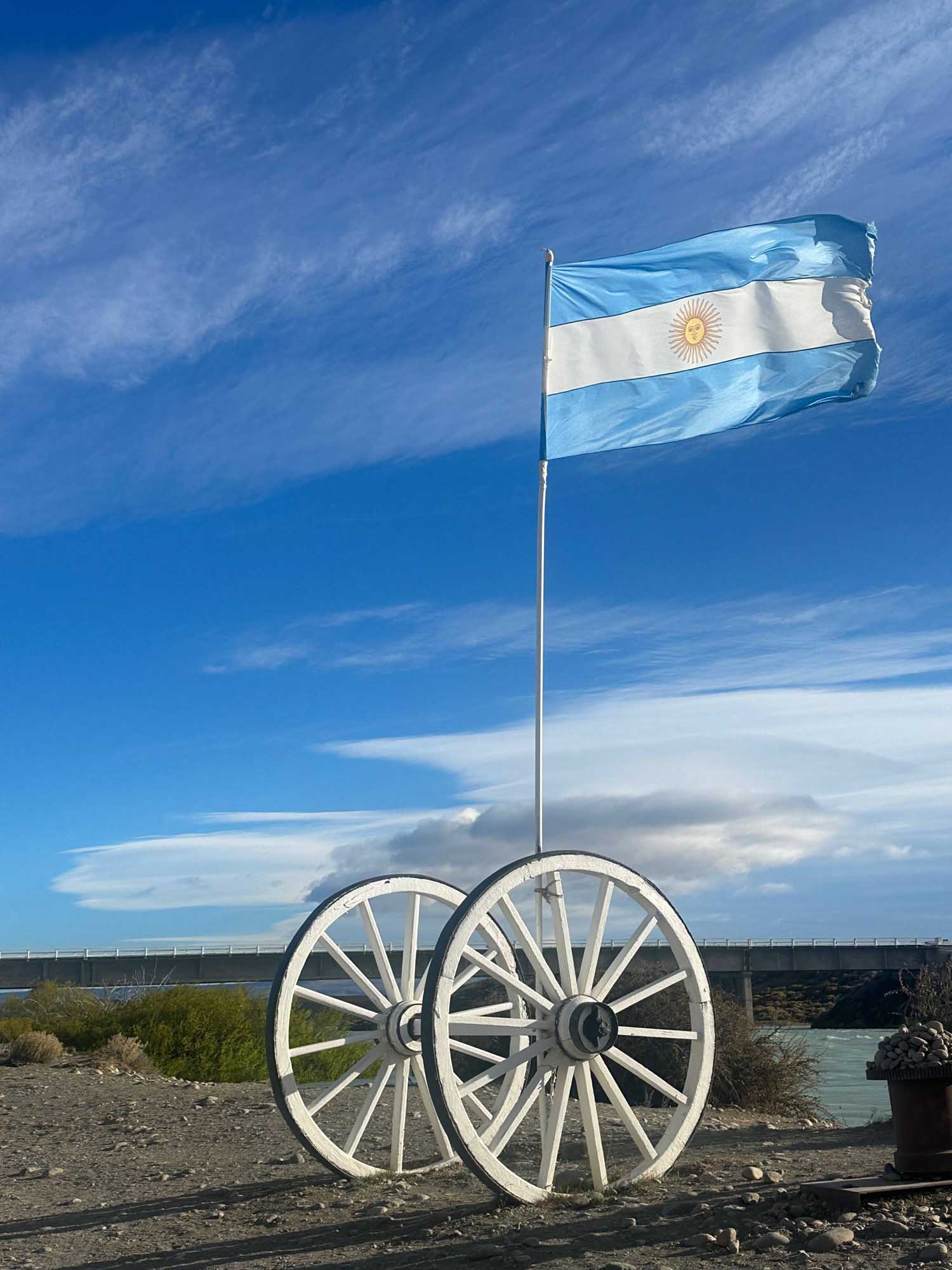 Argentine Flag on Historic Wagon The Argentine flag flies proudly from a flagpole mounted on a pair of white wooden wagon wheels against a vibrant blue sky with wispy clouds. The scene captures a historical monument or display near a waterway and bridge, likely in southern Argentina, representing the region's pioneering heritage.