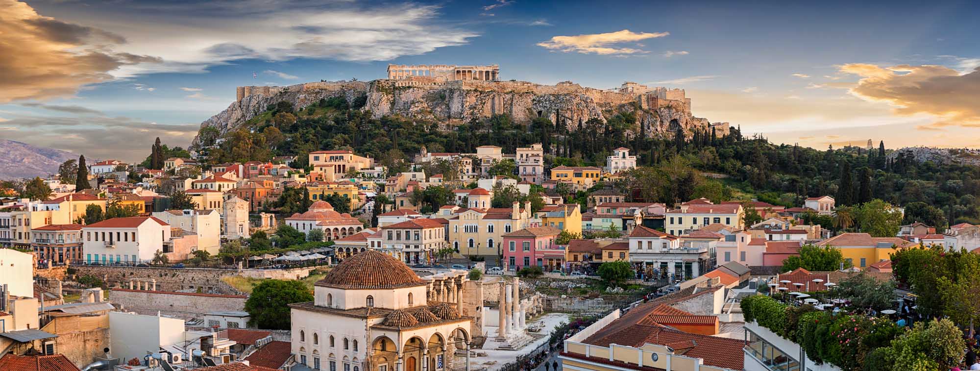 A panoramic view of Athens' historic old town with colorful neoclassical buildings in the foreground and the illuminated Parthenon Temple atop the Acropolis in the background, captured during golden hour sunset with dramatic clouds overhead.