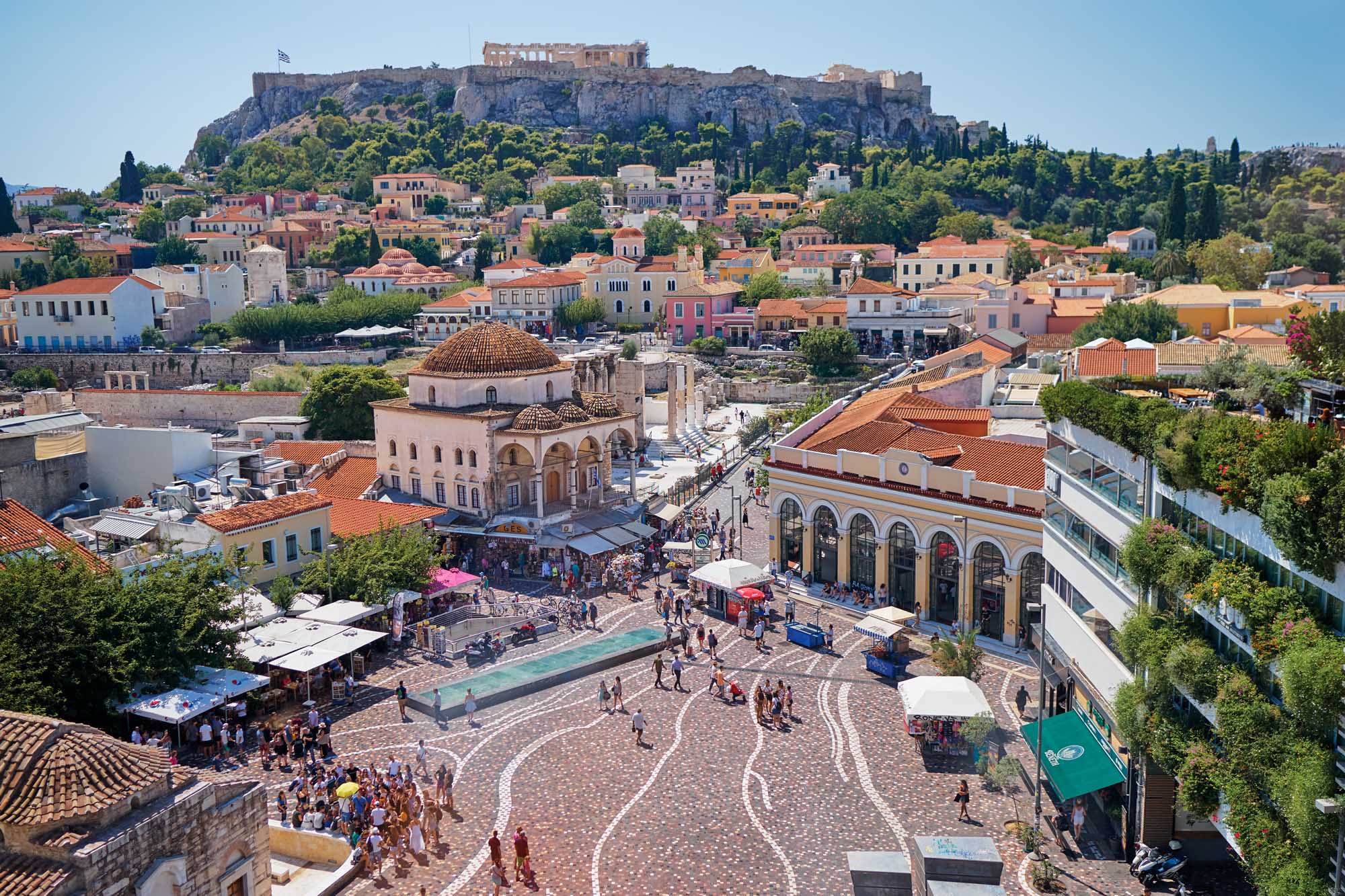 A vibrant daytime aerial view of Athens' historic Monastiraki Square featuring the distinctive domed mosque, colorful neoclassical buildings, bustling marketplace, and the iconic Acropolis perched dramatically on the hilltop in the background under clear blue skies.