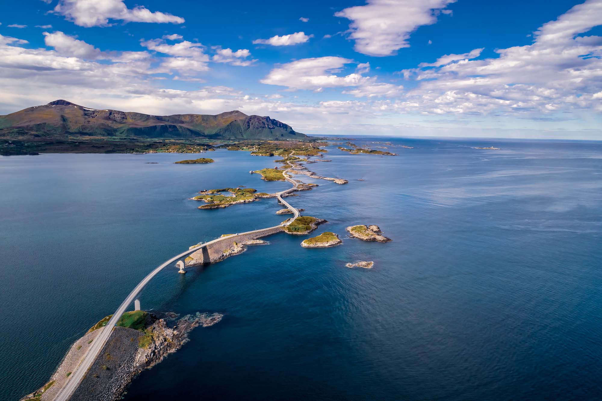 An aerial photograph of the Atlantic Ocean Road (Atlanterhavsveien) in Norway, showcasing a dramatic serpentine causeway connecting small islands across the blue Atlantic waters. The road winds through a stunning coastal landscape with mountains visible in the background and pristine blue skies dotted with white clouds.