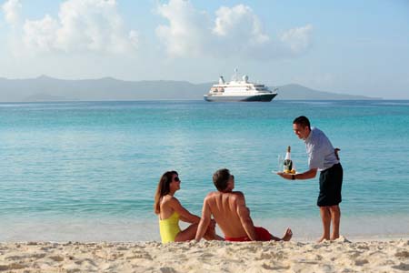 A family enjoys a tropical beach vacation with a man serving drinks to two children sitting in the sand, while a large cruise ship anchors in the turquoise waters offshore with mountainous islands visible in the distant background.