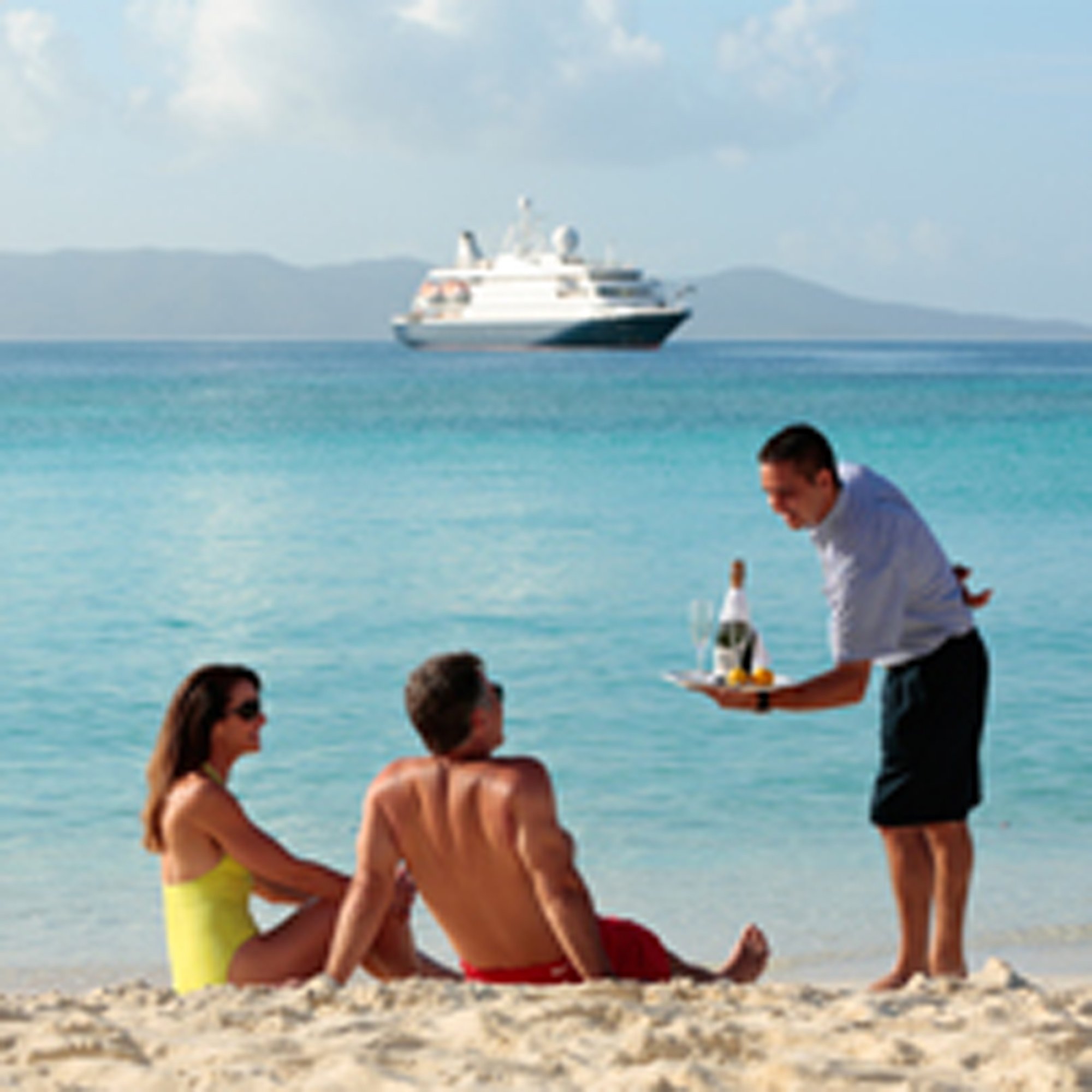 A waiter serves refreshments to beachgoers on a pristine tropical beach while a large cruise ship anchors in the turquoise waters offshore. The scene captures the luxury and convenience of cruise ship dining with beach service during a port stop.