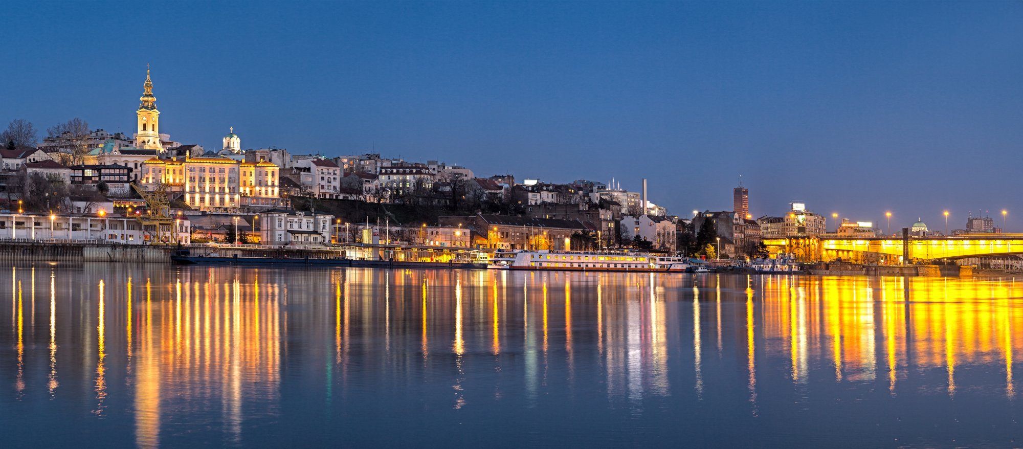 Belgrade Waterfront at Twilight A stunning twilight view of Belgrade's Danube riverfront, featuring illuminated historic architecture reflected in calm waters. The golden-lit buildings, distinctive church spires, and riverside promenade showcase the city's architectural charm and vibrant nightlife along the Danube River.