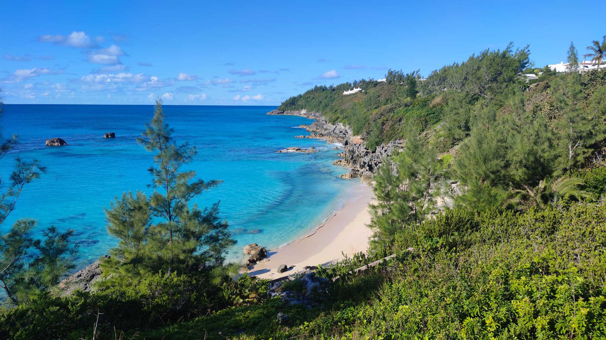 A pristine tropical beach cove nestled between lush green cliffside vegetation and turquoise Atlantic waters, featuring pink-hued sand, rocky outcrops, and whitewashed colonial architecture visible on the hilltop above.