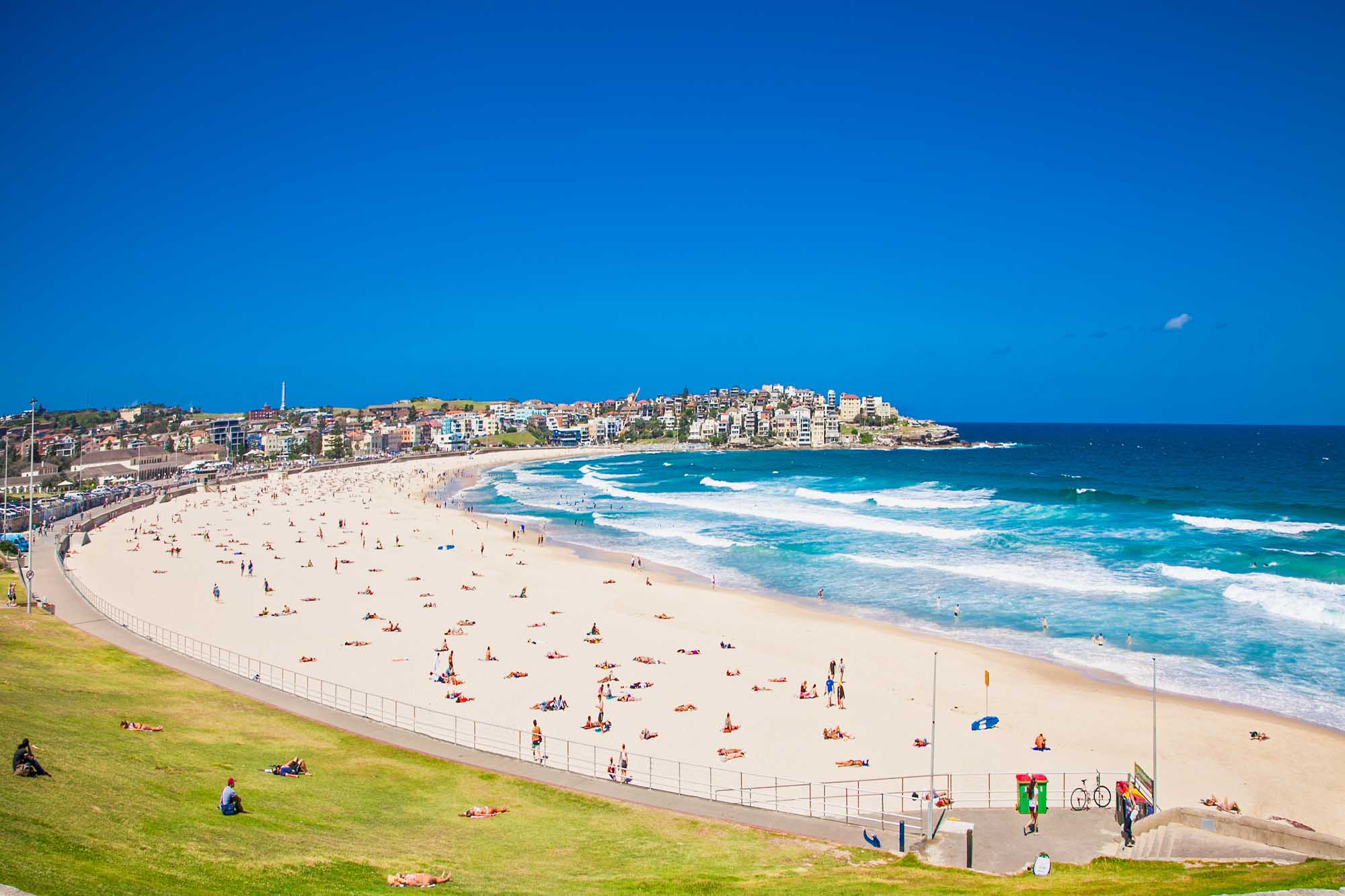 Bondi Beach Sydney on a Sunny Day A vibrant aerial view of Bondi Beach in Sydney, Australia, featuring golden sand packed with beachgoers, turquoise ocean waves, and the iconic beachside suburb with colorful residential buildings cascading up the hillside under a brilliant blue sky.