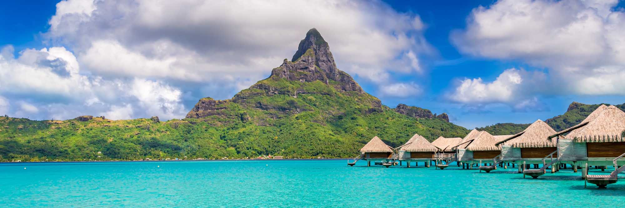 A stunning tropical paradise view of Bora Bora Island in French Polynesia, featuring iconic thatched-roof overwater bungalows in turquoise lagoon waters, with the dramatic volcanic Mount Otemanu peak rising majestically in the background under a bright blue sky with white clouds.
