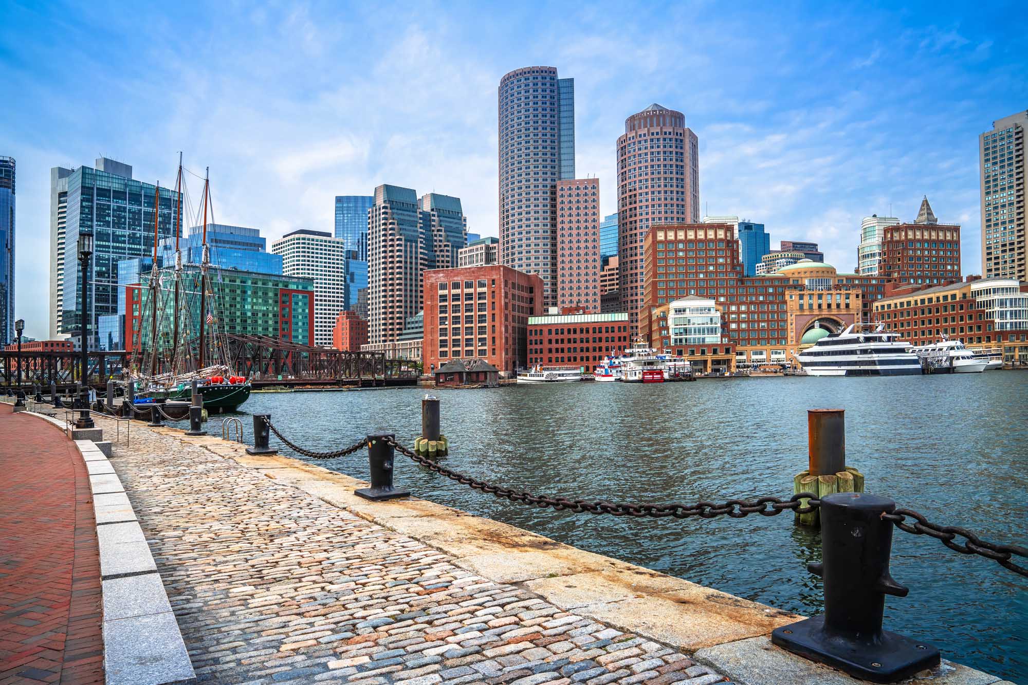Boston Harbor Waterfront with Historic Skyline A picturesque view of Boston's waterfront district featuring the iconic cityscape with a mix of historic red-brick buildings and modern glass skyscrapers. The image showcases moored sailing vessels and cruise ships at the pier, with a clear blue sky and calm harbor waters reflecting the vibrant urban landscape.