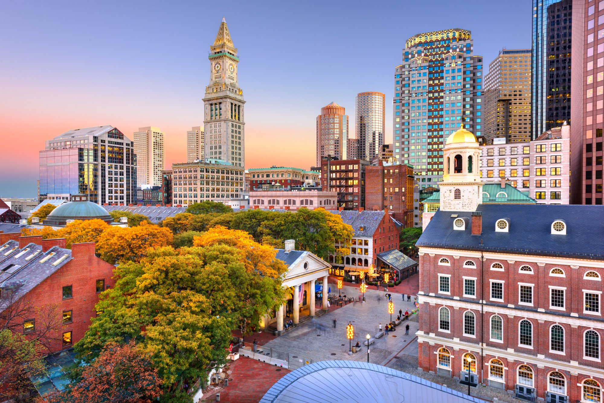A stunning autumn view of Boston's iconic skyline featuring the historic Faneuil Hall with its distinctive cupola in the foreground, surrounded by vibrant fall foliage, colonial-era brick buildings, and modern glass towers illuminated by the golden light of sunset.