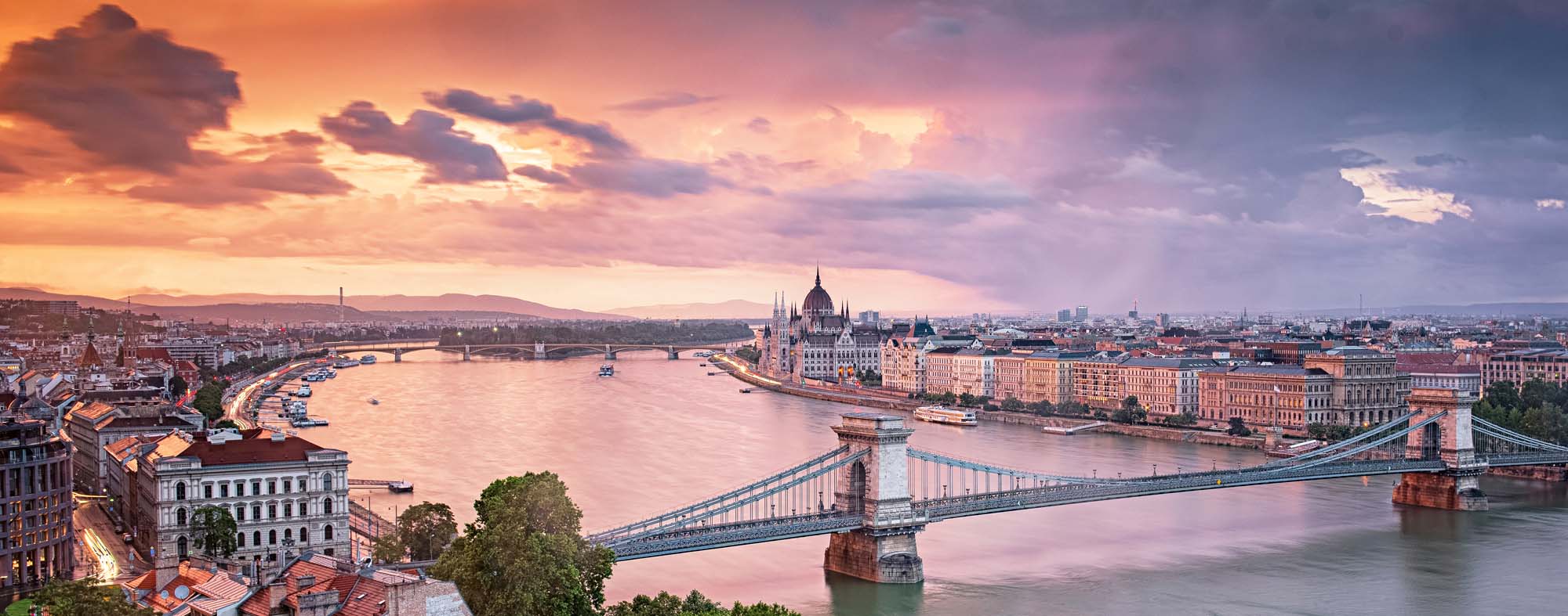 A stunning panoramic view of Budapest at golden hour, featuring the iconic Chain Bridge spanning the Danube River with the Hungarian Parliament Building illuminated on the right bank. The cityscape is bathed in warm orange and pink hues as the sun sets behind dramatic clouds, reflecting beautifully across the water.