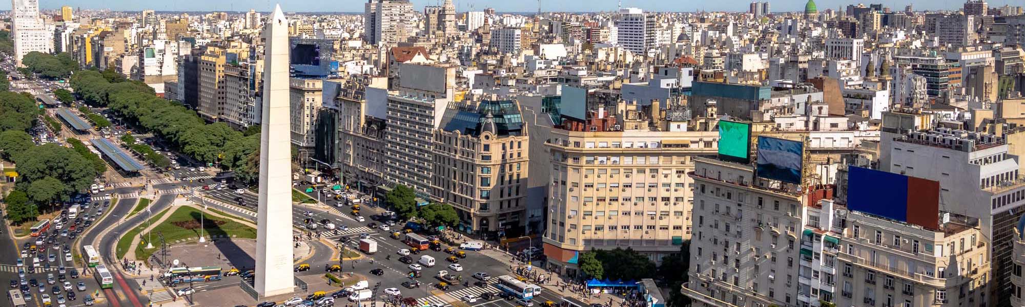 Buenos Aires Downtown Cityscape with Obelisk An expansive aerial view of Buenos Aires' iconic downtown district featuring the prominent Obelisco monument at the center of Avenida 9 de Julio. The image showcases the bustling urban landscape with characteristic belle epoque architecture, busy streets filled with vehicles and pedestrians, lush green parks, and the dense cityscape extending to the horizon.