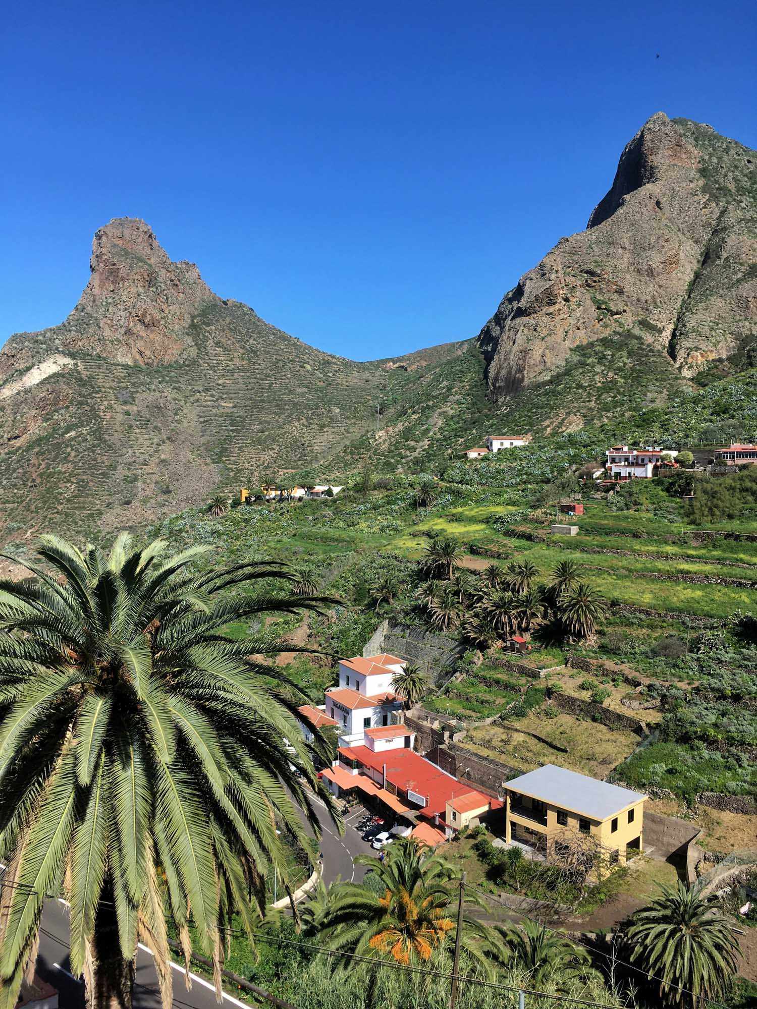 A picturesque valley settlement nestled between dramatic volcanic peaks in the Canary Islands, featuring traditional white and colorful buildings with terracotta roofs, lush green agricultural terraces, palm trees, and a clear blue sky. The landscape showcases the unique combination of arid mountainous terrain and verdant valley cultivation typical of the archipelago's diverse geography.