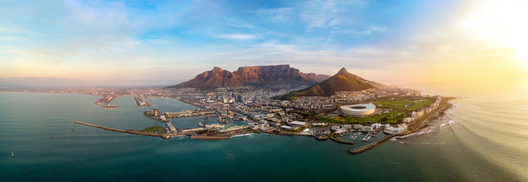 Cape Town Aerial View with Table Mountain An expansive aerial photograph of Cape Town, South Africa at sunset, showcasing Table Mountain dominating the cityscape, the iconic white oval stadium in the foreground, pristine turquoise waters of Table Bay, and the surrounding urban landscape bathed in golden light.