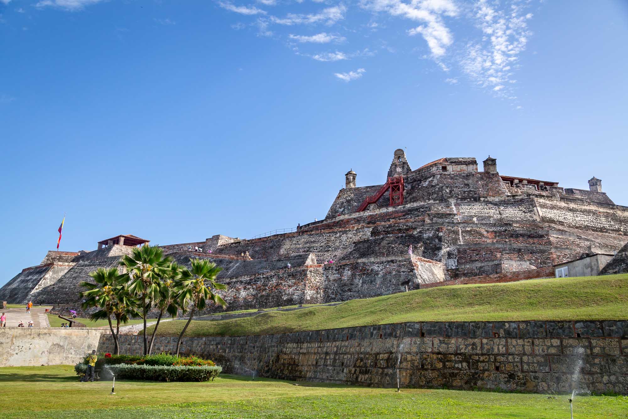 A UNESCO World Heritage Site fortress featuring distinctive red brick and stone ramparts with palm trees in the foreground, located in Cartagena, Colombia. The imposing 16th-17th century military structure showcases Spanish colonial architecture with its characteristic angled bastions and weathered fortification walls.