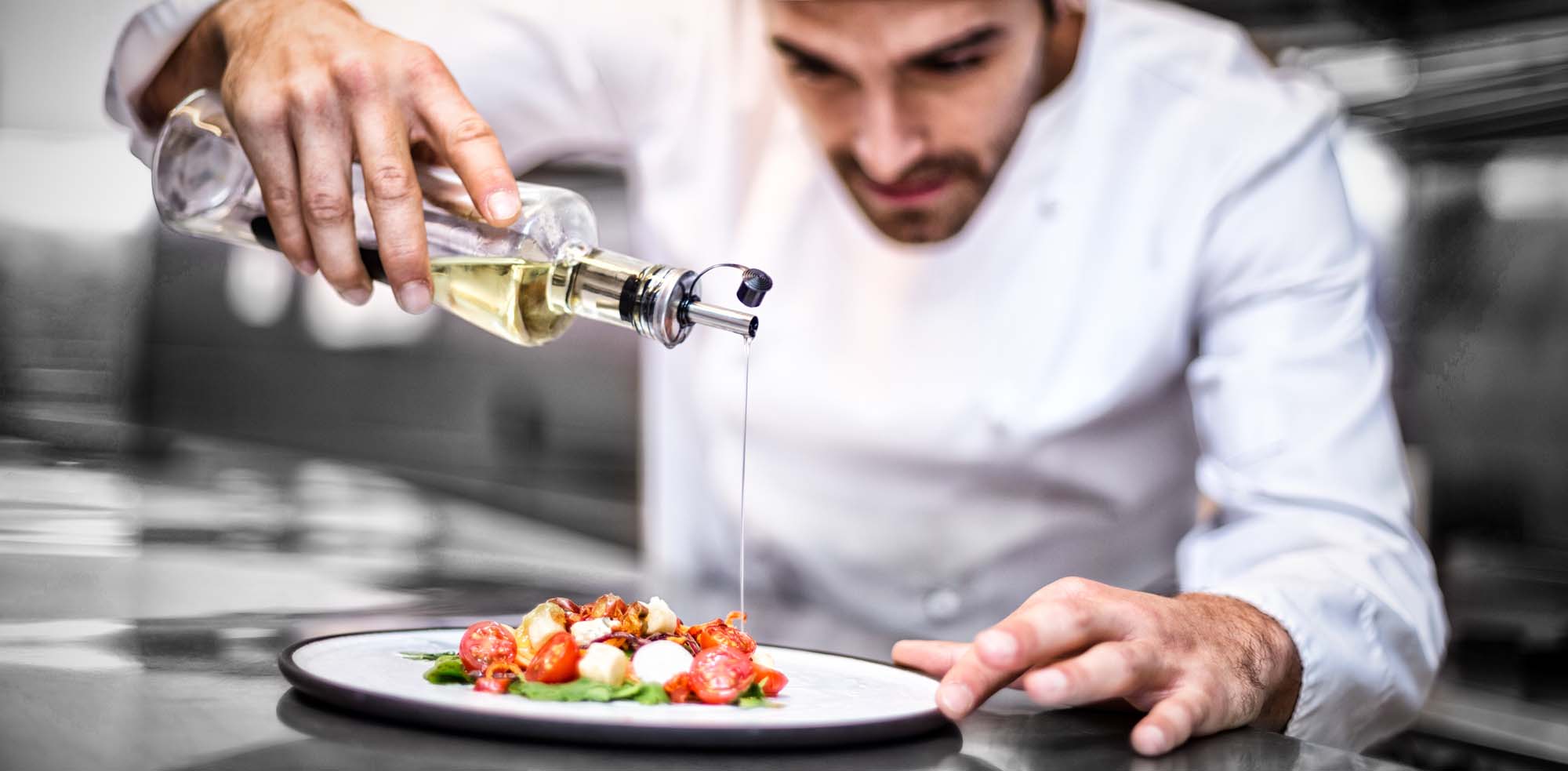 Chef Drizzling Olive Oil on Plated Dish A professional chef in a white chef's coat carefully drizzles golden olive oil over an artfully plated dish featuring sliced tomatoes and other fresh ingredients in a commercial kitchen setting. The image captures a moment of culinary precision and fine dining preparation.