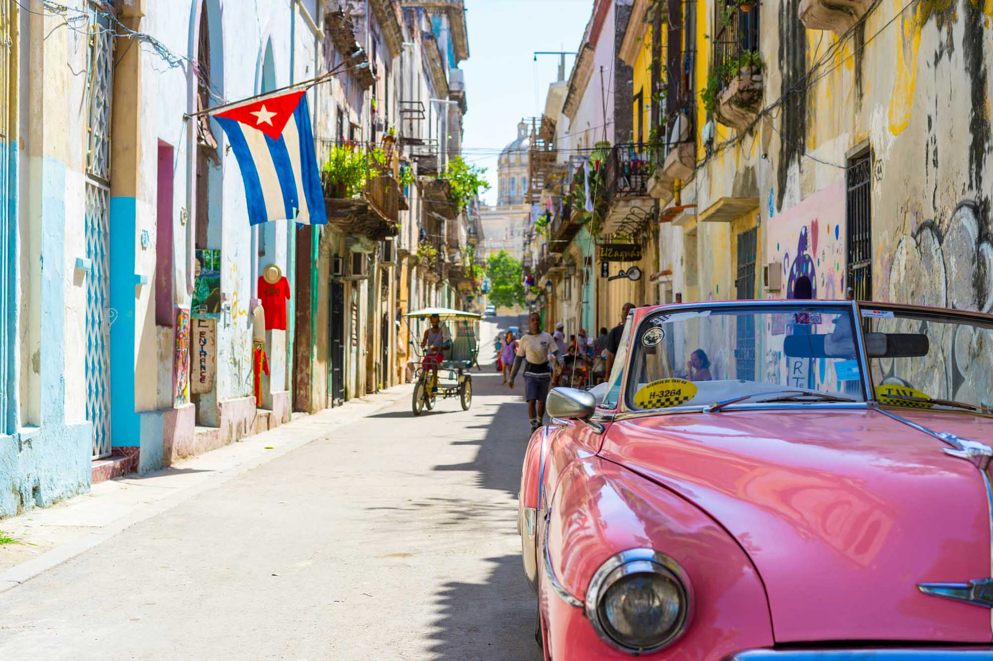 A vibrant street scene in Havana, Cuba featuring a pink vintage American car parked along a colorful colonial building, with a Cuban flag hanging overhead. The image captures the iconic charm of Old Havana with its weathered architecture, narrow cobblestone streets, traditional horse-drawn cart, and the distinctive aesthetic that makes Cuba a unique Caribbean destination.