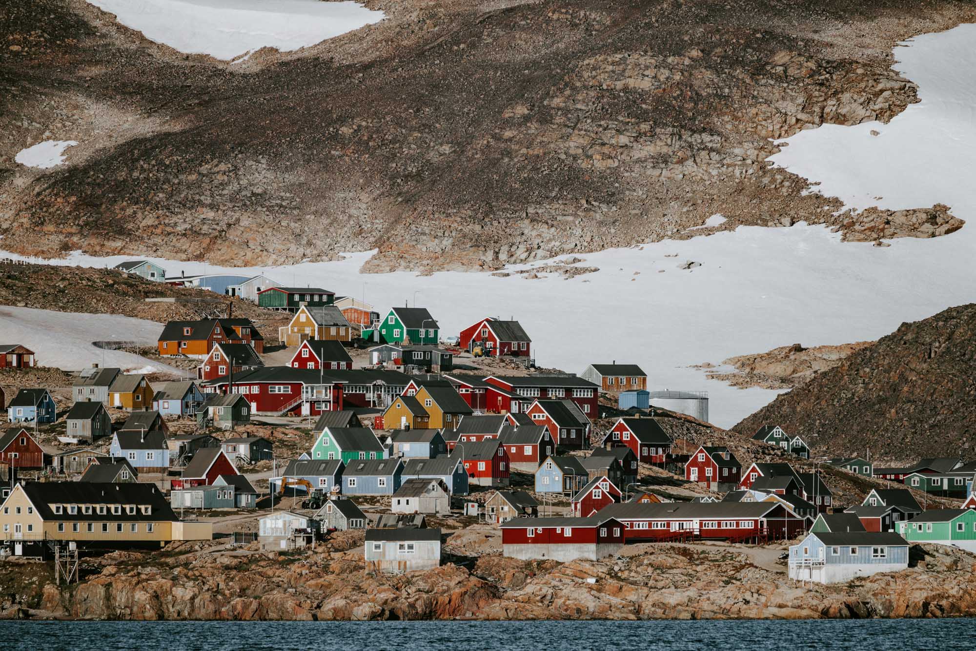 Colorful Arctic Village with Snowy Mountains A vibrant Arctic settlement featuring densely packed houses painted in traditional bright colors—reds, greens, yellows, and blues—nestled against stark rocky terrain and patches of snow. The picturesque village sits along the waterfront, characteristic of Greenlandic settlements with their distinctive architectural style and palette.