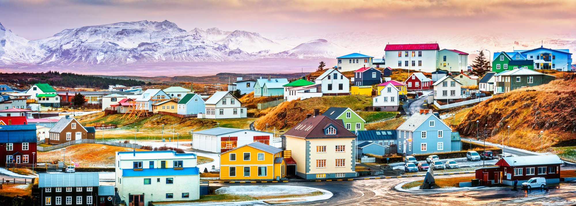 A vibrant Greenlandic settlement featuring distinctive brightly-colored houses with grass-covered roofs arranged across rolling tundra, with a snow-capped mountain range dominating the background and small fishing boats visible in the harbor. The scene captures the unique Arctic architectural style and natural landscape characteristic of Greenland's remote communities.