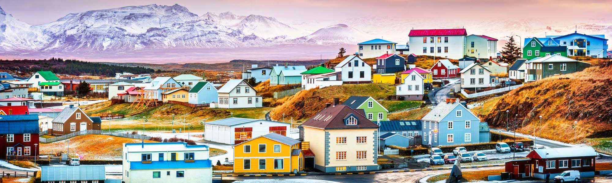 A vibrant Arctic coastal settlement featuring distinctively colorful houses in bright hues of blue, yellow, red, and green nestled against snow-capped mountains and moorland terrain. The picturesque village showcases traditional Nordic architecture with a mix of residential buildings, small boats, and vehicles arranged along a waterfront, characteristic of Greenlandic settlements.