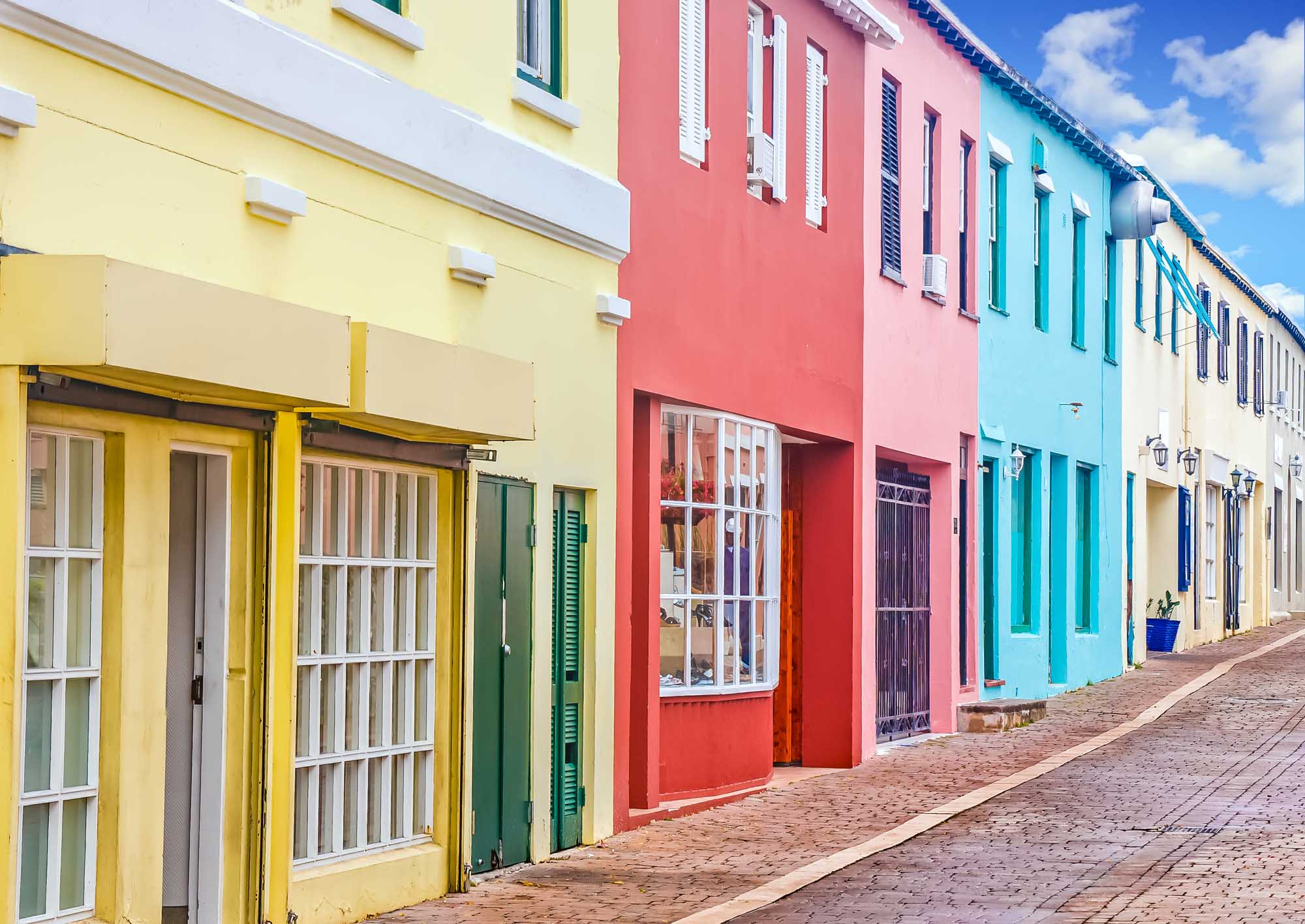 A picturesque street lined with vibrant pastel-colored colonial buildings featuring yellow, pink, turquoise, and cream facades. White shuttered windows and traditional wooden doors characterize the historic architecture, with a brick-paved street leading through this charming residential or commercial district typical of Bermuda's distinctive aesthetic.