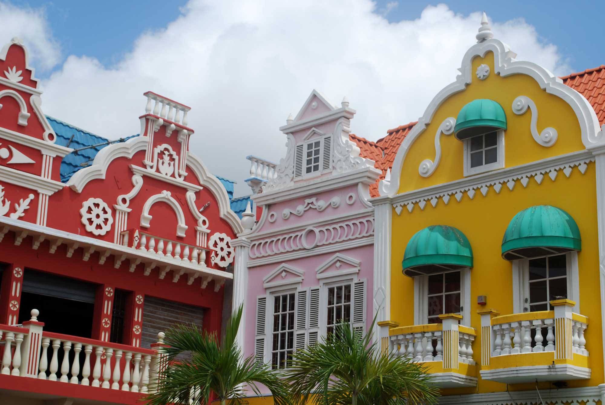 A vibrant row of Dutch Colonial buildings featuring bold primary colors—red, pink, and yellow—with white ornamental trim, arched windows, and turquoise awnings. The iconic architecture reflects Caribbean colonial heritage with distinctive peaked roofs and decorative balustrades, typical of Curaçao's historic waterfront district.