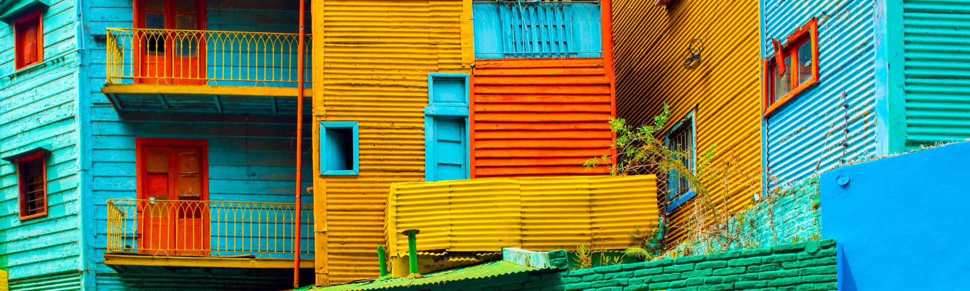 Colorful Colonial Buildings in La Boca A vibrant street scene featuring iconic brightly painted corrugated metal buildings in shades of turquoise, yellow, orange, and red with decorative iron railings and shuttered windows. This picturesque neighborhood showcases the distinctive architectural style and rich cultural heritage of Buenos Aires' historic La Boca district.