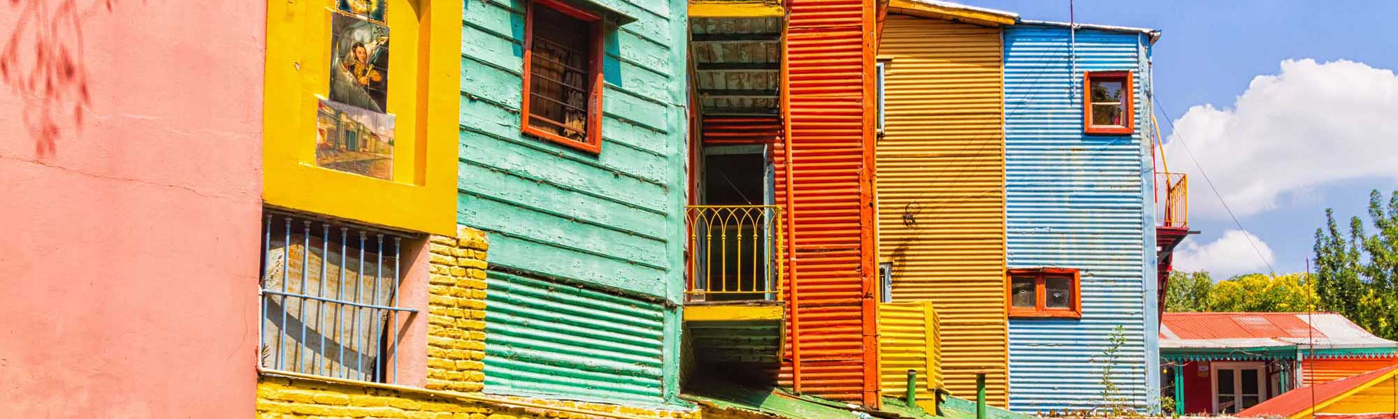 Colorful Historic Buildings in La Boca A vibrant row of traditionally painted corrugated metal houses in bright primary colors including pink, yellow, green, red, and blue. These iconic structures feature characteristic architectural details with shuttered windows, metal railings, and corrugated siding typical of the historic La Boca neighborhood.