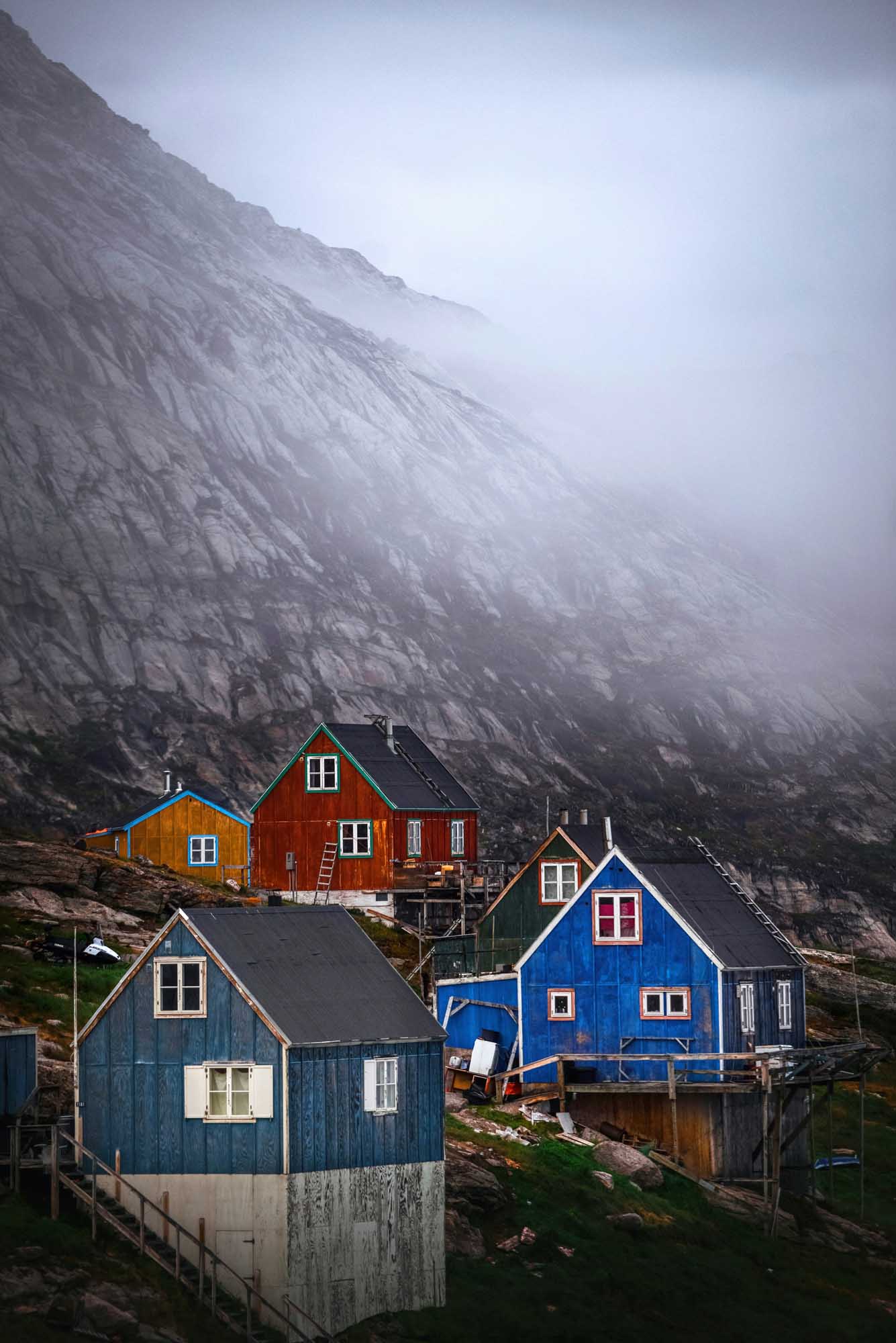 Colorful Houses Beneath Misty Arctic Cliffs A picturesque collection of brightly colored traditional houses nestled at the base of a towering, mist-shrouded rocky cliff in the Arctic. The distinctive architecture features vibrant blue, red, and yellow wooden dwellings with white-trimmed windows, characteristic of Greenlandic settlements, creating a striking contrast against the dramatic gray mountainous landscape.