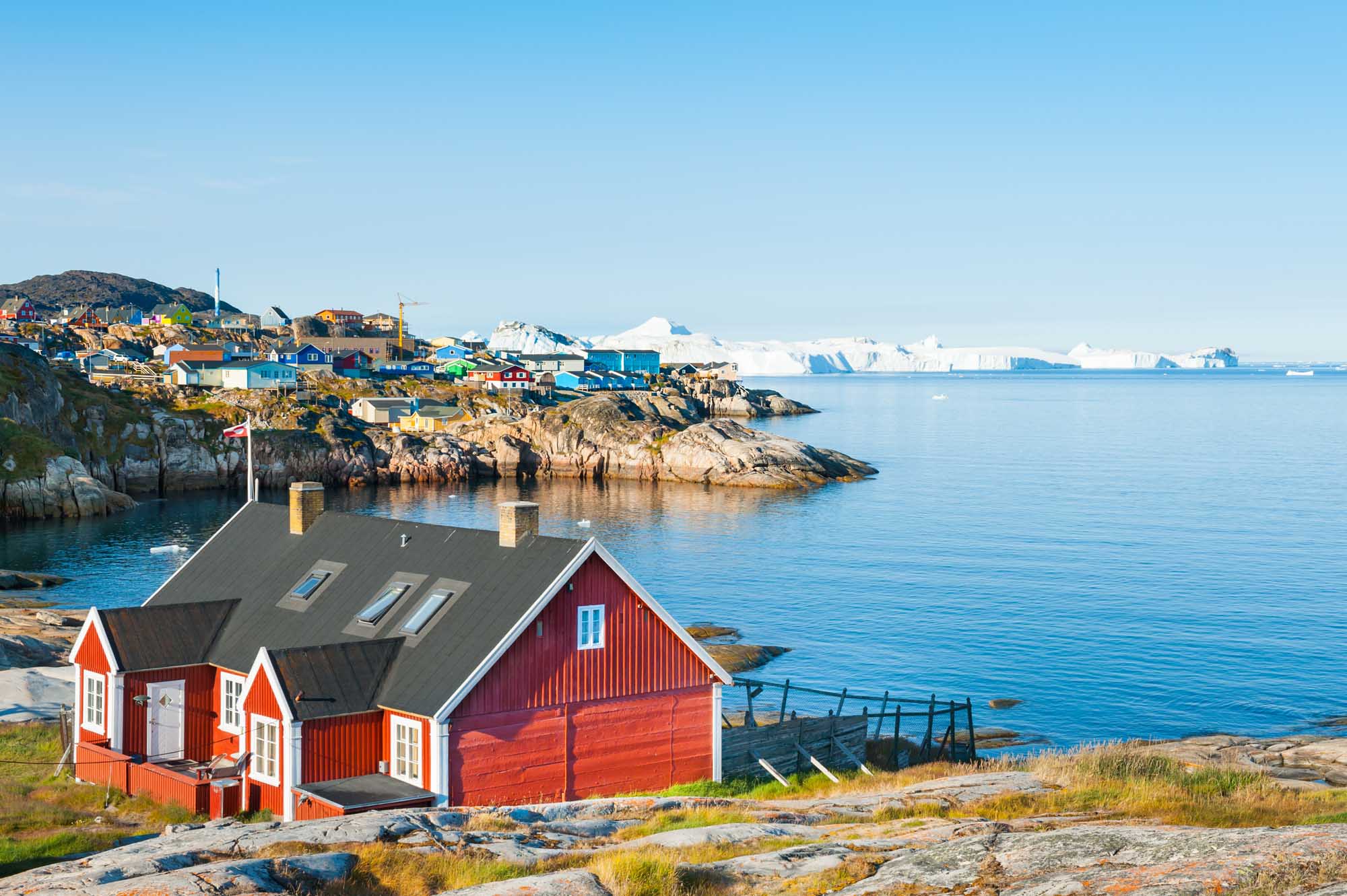 Colorful Houses in Ilulissat, Greenland A picturesque red Greenlandic house sits on rocky terrain overlooking the Atlantic Ocean, with the vibrant settlement of Ilulissat visible across the bay and icebergs dotting the horizon. The scene captures the distinctive Arctic architecture and pristine coastal landscape characteristic of western Greenland.