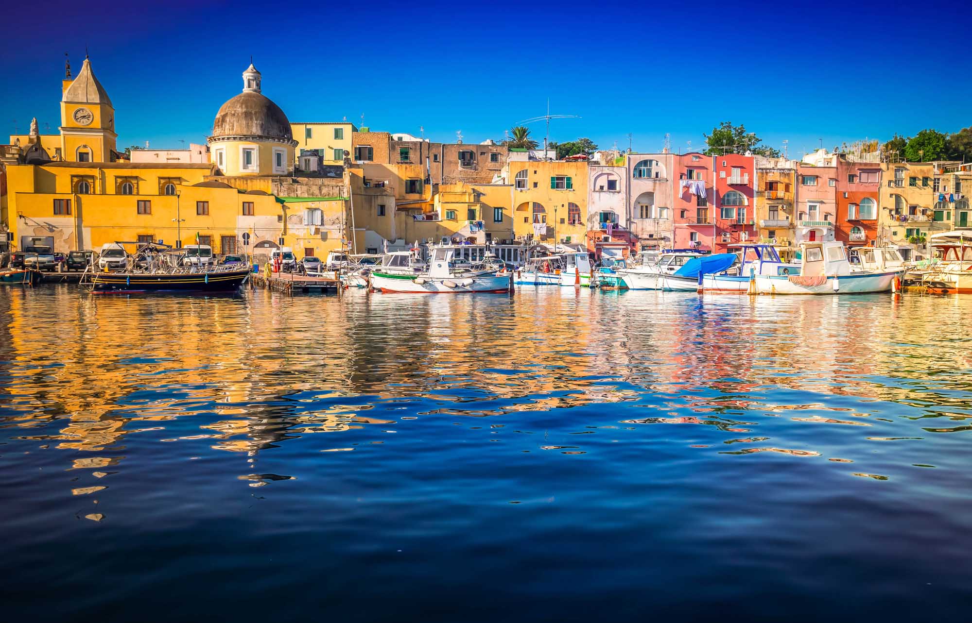 A vibrant Mediterranean harbor scene featuring the iconic Marina Grande of Procida Island, with densely packed pastel-colored buildings in yellow, pink, orange, and cream tones reflecting in the calm blue waters. Traditional fishing boats are moored along the waterfront, with a prominent domed church visible among the historic architecture.