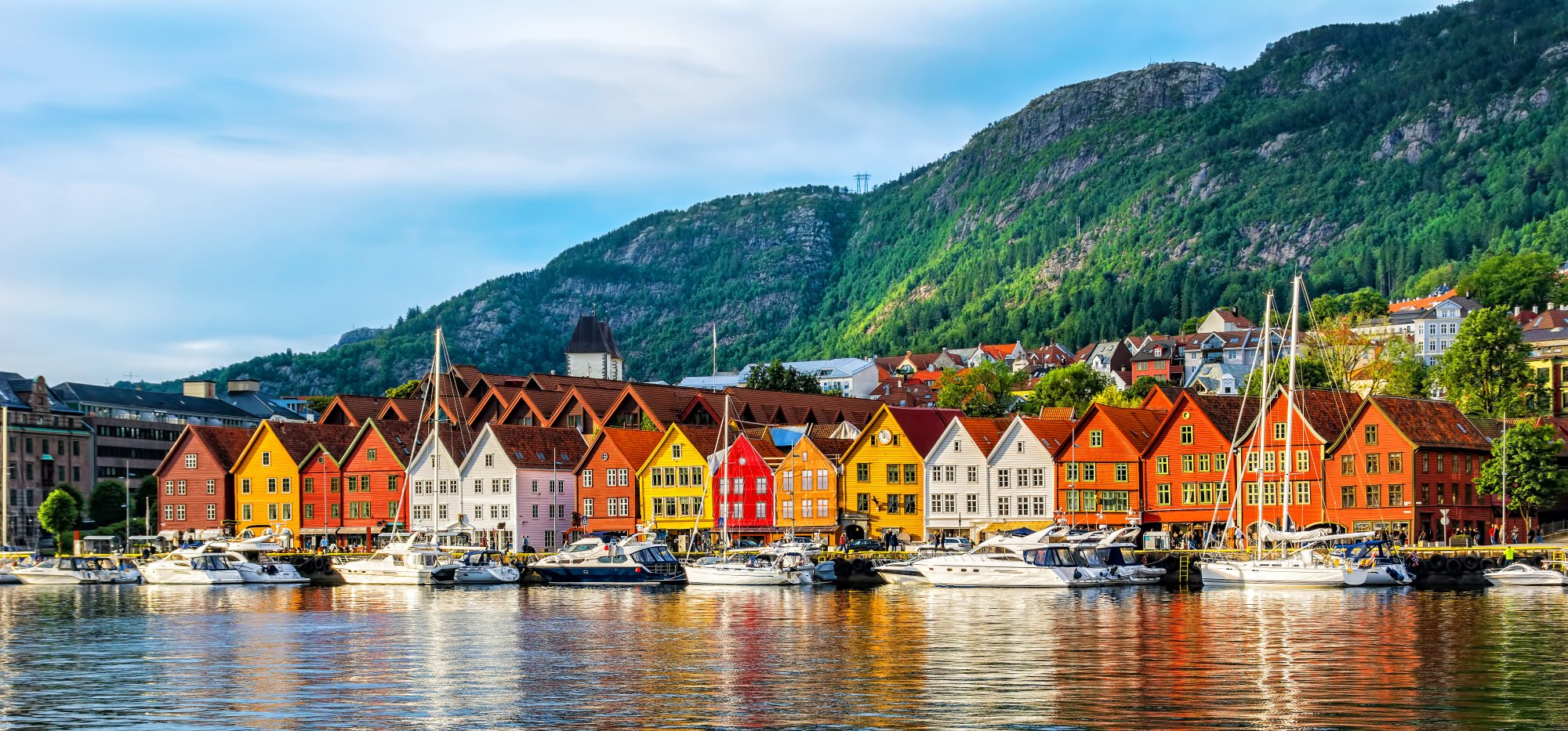Colorful Waterfront Houses in Bergen, Norway A picturesque Scandinavian waterfront scene featuring a row of vibrant traditional wooden houses in shades of red, orange, yellow, and white, with moored sailboats in the foreground and dramatic forested mountains rising in the background under a clear blue sky.