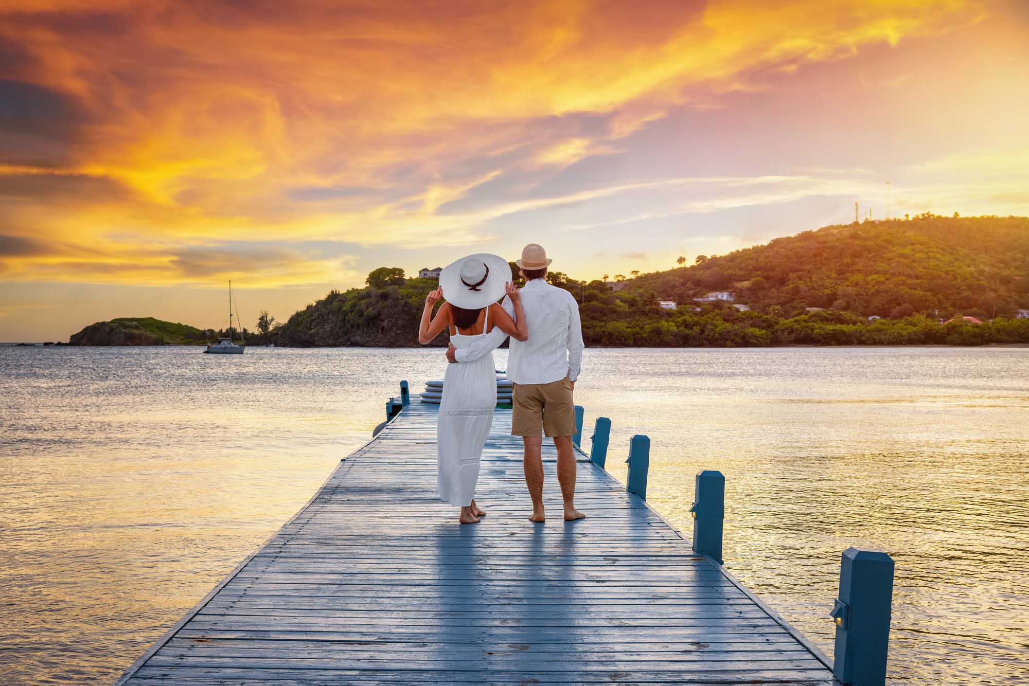 A romantic couple stands on a wooden pier overlooking calm tropical waters at golden sunset, with lush green islands and sailboats visible in the background. The woman wears a white flowing dress and wide-brimmed hat while the man wears light casual clothing, capturing an idyllic honeymoon or vacation moment in a Caribbean paradise setting.
